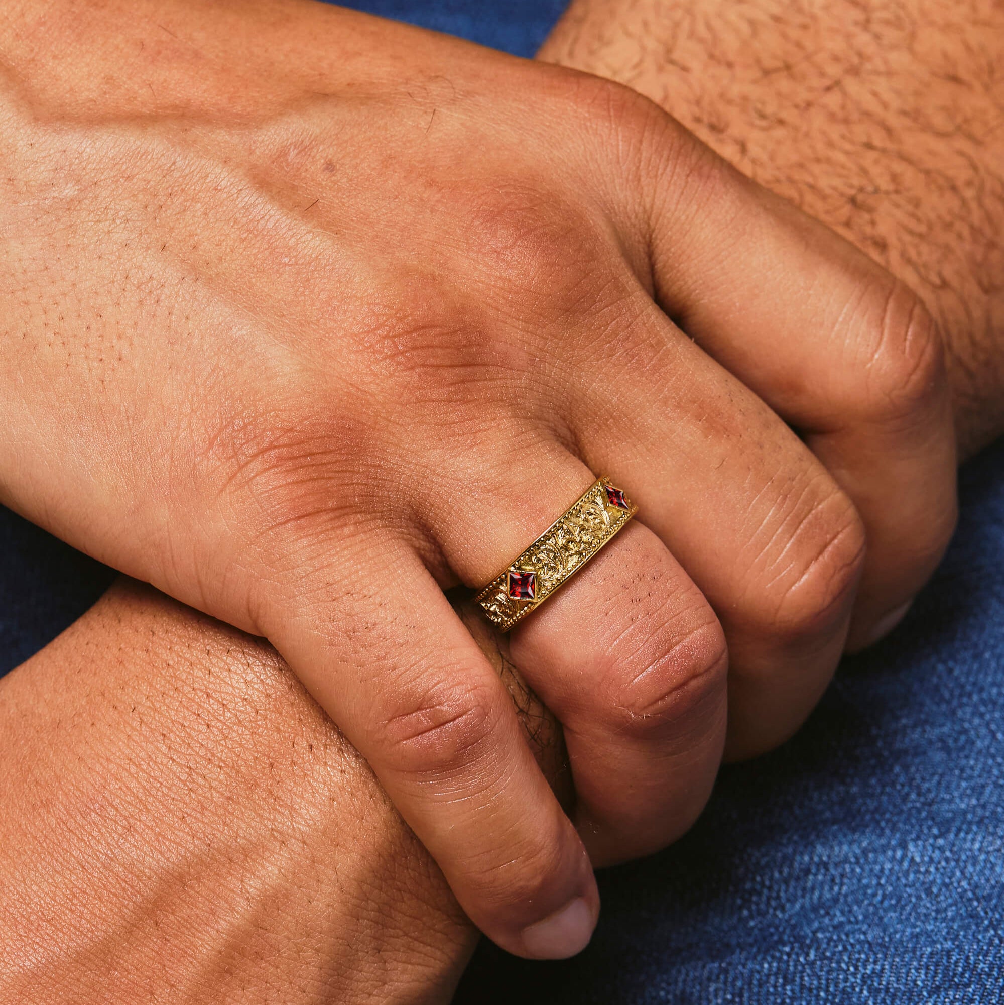 Close-up of a hand wearing a gold ring with red stones on a blue fabric background